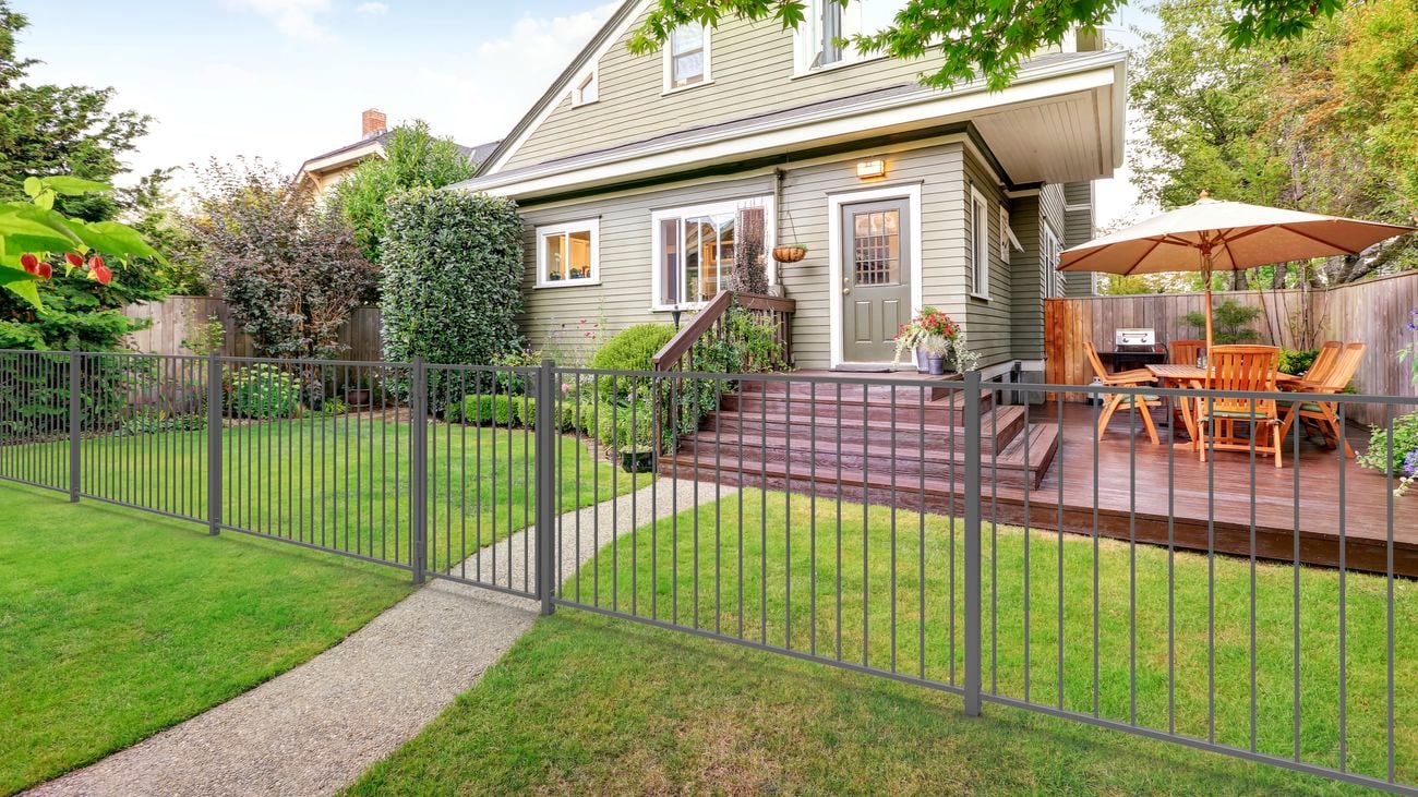Grey fence sits in the front yard of a house, with a wooden deck on the side 