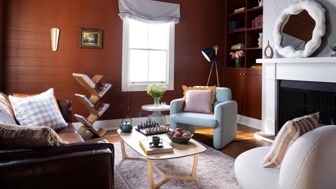 Living room setting with three armchairs, maroon feature wall, fire place, decorative mirror and bookshelf.