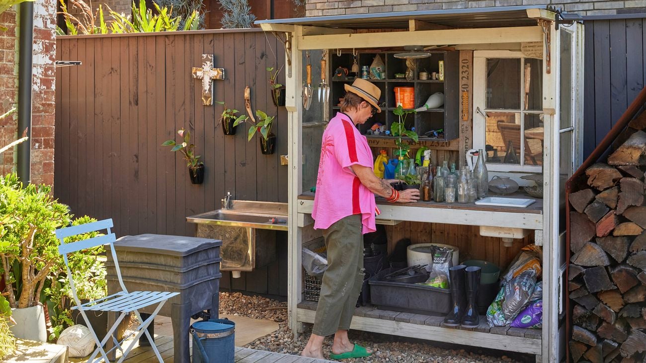 Penny wearing a hat and pink shirt stands at an outdoor potting bench with shelves of jars, pots and garden tools. Backyard scene with timber wall, small bistro chair and table on the deck, a stack of firewood to the right, and boots and supplies under the bench.