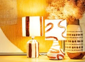 Two abstract paper mache lamps with lights on next to a vase on a table in front of an artwork and brick wall