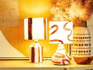 Two abstract paper mache lamps with lights on next to a vase on a table in front of an artwork and brick wall