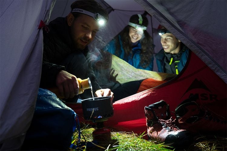 Three people in a tent wearing head torches cooking dinner