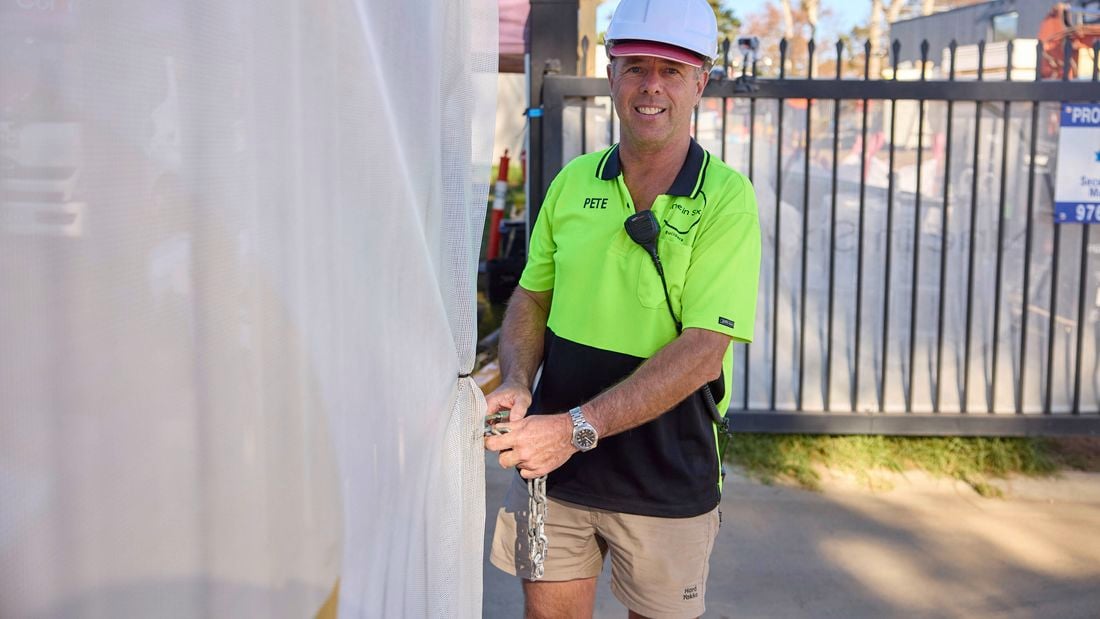 A man in a yellow hi-vis t-shirt and white hardhat looks at the camera while holding a lock chain beside a fence on a construction site