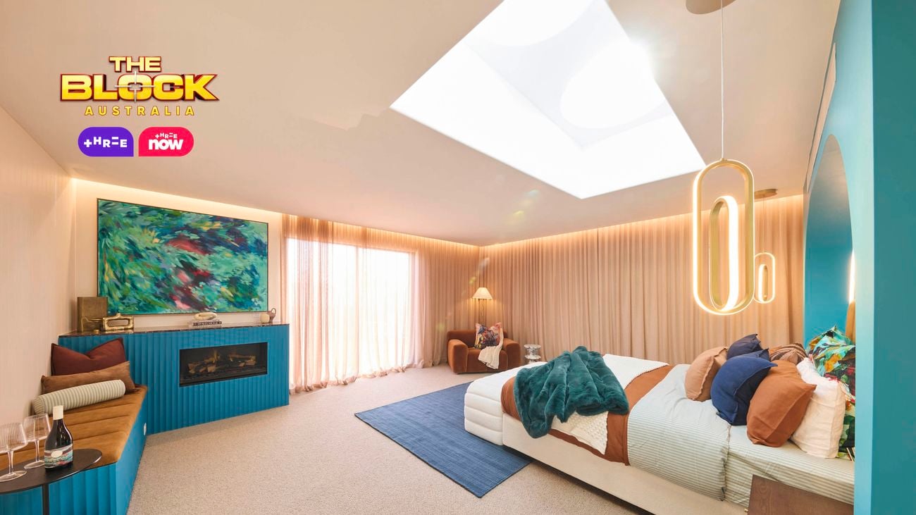 A modern bedroom featuring a large bed with white and brown bedding, colorful pillows, and a green throw blanket. The blue headboard wall has an arched design and pendant lights on either side. A wooden nightstand with decorative items sits next to the bed. The room includes a blue rug, beige curtains, and a rectangular skylight in the ceiling. 