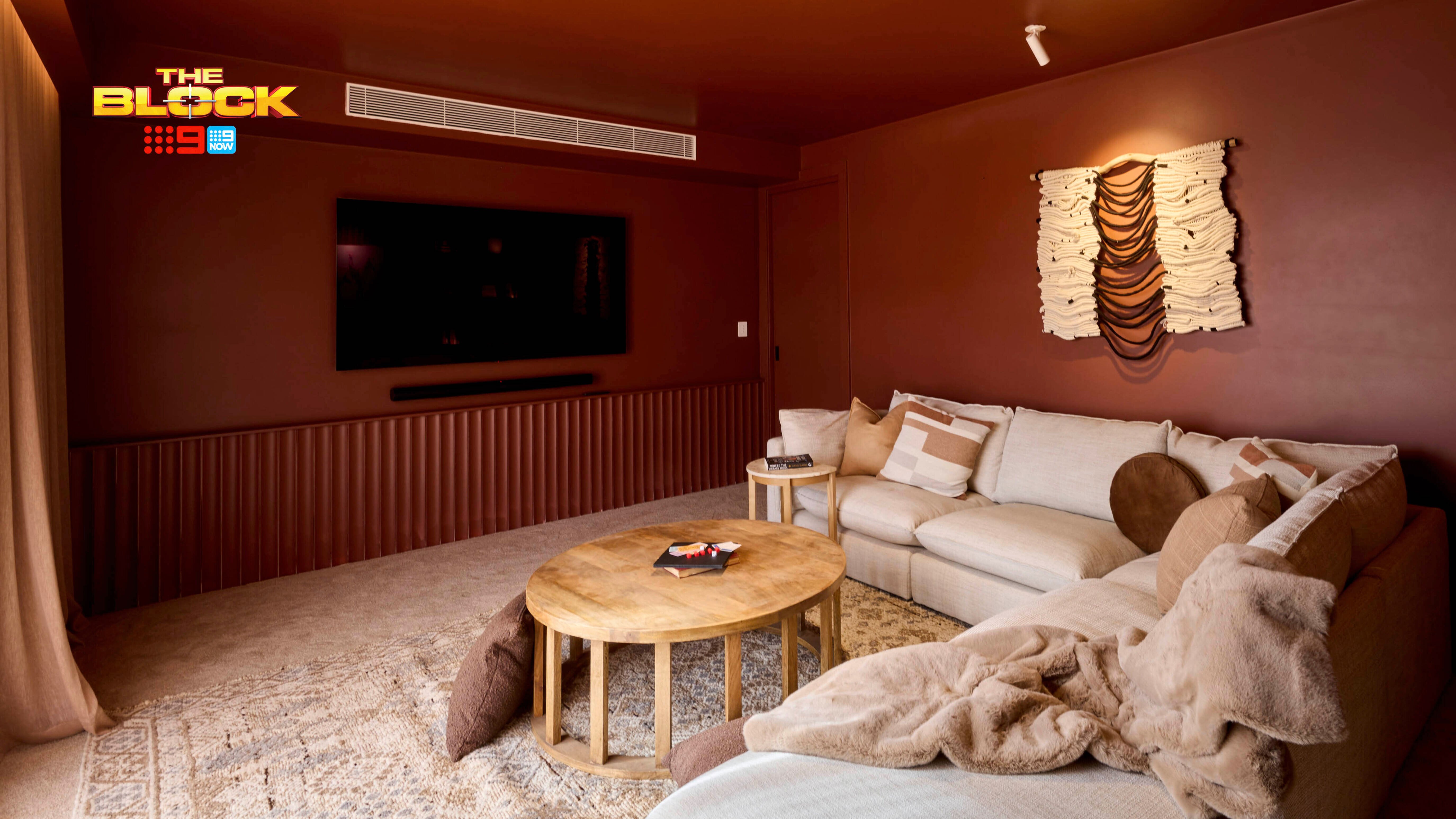 A warm-toned living room featuring a large beige sectional sofa with assorted pillows and throws. A round wooden coffee table sits in front of the sofa, facing a wall-mounted flat-screen TV. The walls are painted brown and decorated with an artistic wall hanging above the sofa. A patterned rug covers the floor.