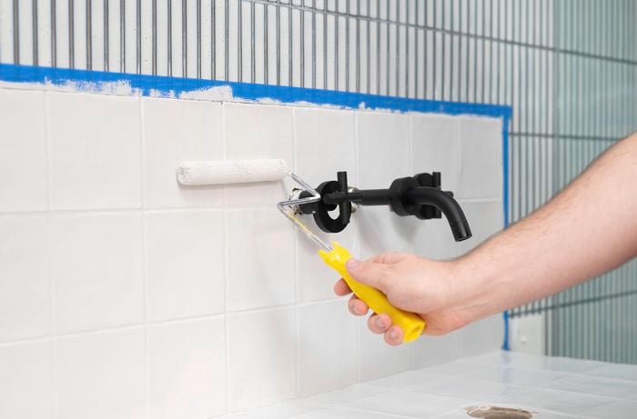 A person is using a small paint roller to apply a second coat of white paint to tiled walls with painter's tape protecting the edges.