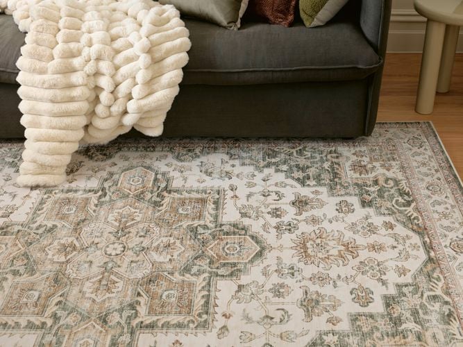 Textured cream throw draped over a dark grey sofa beside a small side table, resting on a faded patterned rug on a wooden floor in a cozy living room setting.