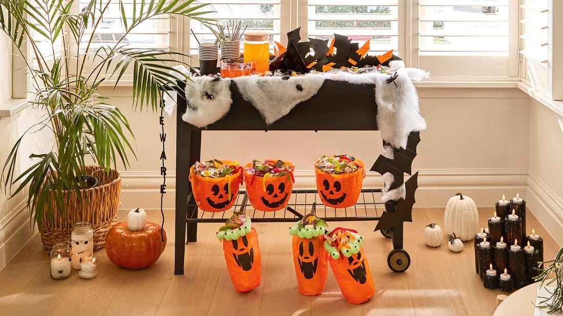 A Halloween craft display by a bay window with a skeleton figure, a decorated trolley holding treats and drinks, hanging bat-themed lanterns, and pumpkin buckets on the floor.