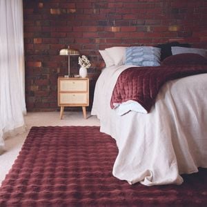 A bedroom with a brick wall, bed, side table with a lamp on it and a burgundy rug on the floor in it.