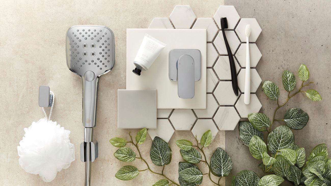 Flatlay image of bathroom fixtures including a stainless steel showerhead, stainless steel shower mixer tap, white hexagon-shaped mosaic tiles and a shower sponge