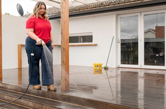 A Bunnings Team Member is watering a wooden deck with a garden hose, while a yellow bucket and a broom can be seen in the background