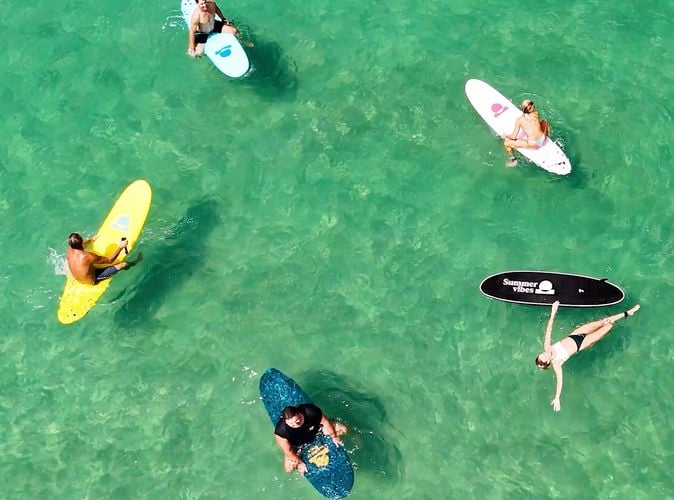 Aerial view of five surfers floating on colourful soft surfboards in clear green water.