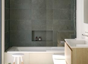 A modern bathroom with grey tiles, a white bathtub with shower, toilet, and a wooden vanity with a white basin. Three green towels hang on a rack to the left next to a white and timber stool.