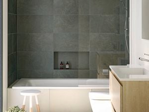 A modern bathroom with grey tiles, a white bathtub with shower, toilet, and a wooden vanity with a white basin. Three green towels hang on a rack to the left next to a white and timber stool.