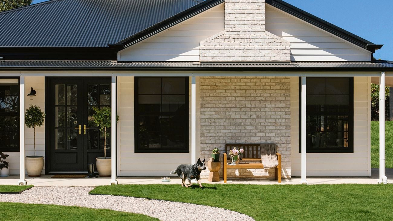 The white farmhouse house at Little Lyndoch Flower Farm with a gravel path through a green lawn, a dog running, and a wooden bench on the porch
