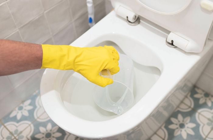 A person wearing a yellow rubber glove is carefully removing excess water from the toilet bowl using a plastic container.
