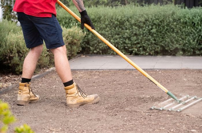 A person is using a rake to loosen the top layer of soil and leveling it, preparing the ground for turf installation. ] 