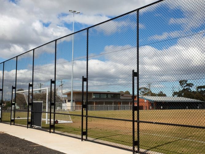 Sports field enclosed by a tall chain-link fence, with goalposts, buildings, and a covered seating area in the background under a partly cloudy sky