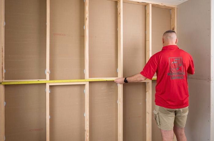 A Bunnings Team Member uses a tape measure to measure a wooden wall frame with vertical beams. 