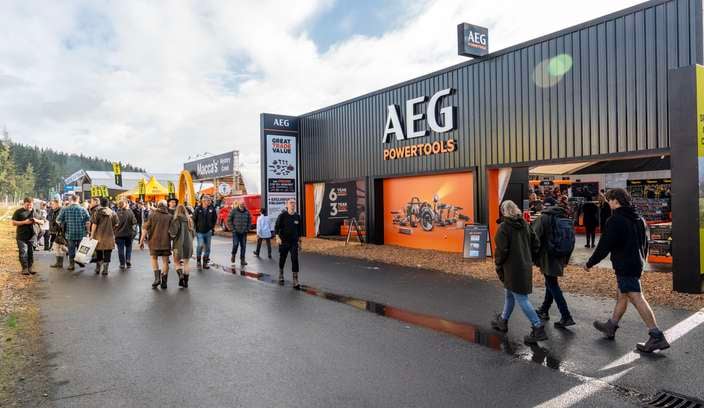 Crowd of people walking along a wet pathway at an outdoor trade event, passing a large black and orange AEG Powertools exhibition stand, with branded signage, display tents, and other vendor booths visible under a partly cloudy sky.