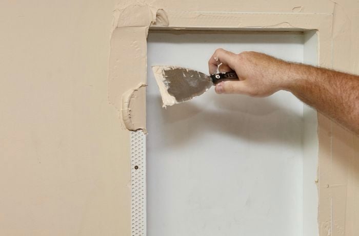 A person applying joint compound around the perforated edge of a niche using a trowel