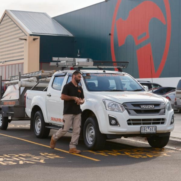 Tradesperson walking beside a white Isuzu utility vehicle towing a trailer outside a Bunnings Warehouse store.