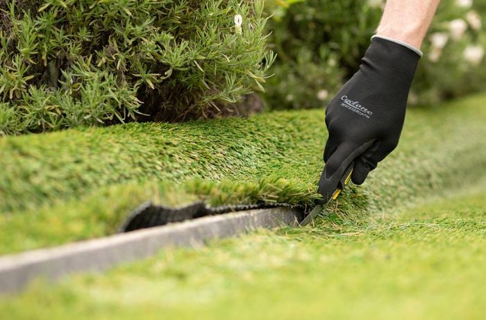A person wearing gloves is cutting the synthetic turf to the desired size using a utility knife.