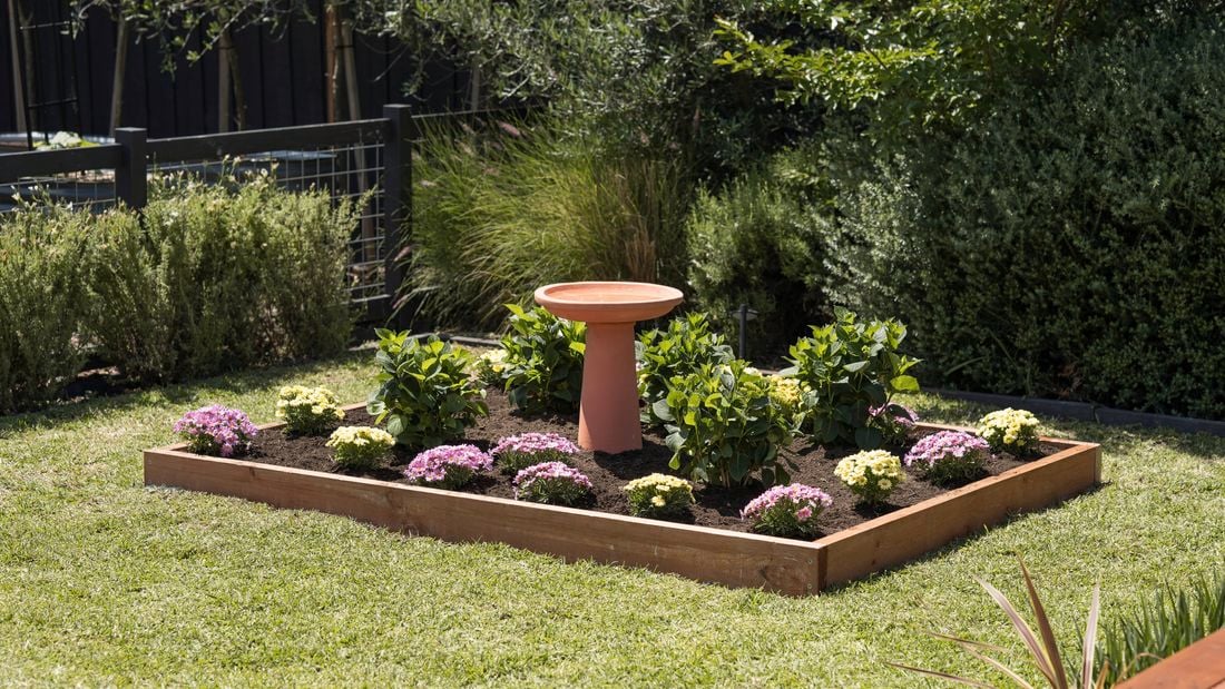 A raised garden bed featuring pink and yellow flowers, green shrubs, and a terracotta birdbath.