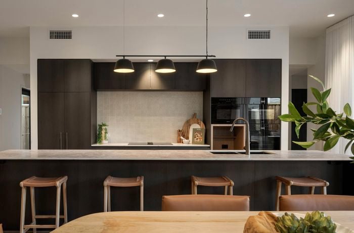 Modern complete installed kitchen with pendant lights, stools, long benchtop, dark cabinetry and appliances.