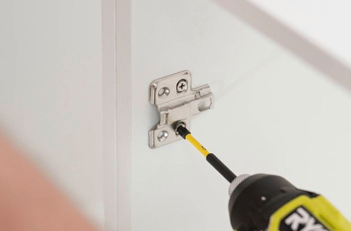 A person uses a power drill to install a hinge plate inside a cabinet