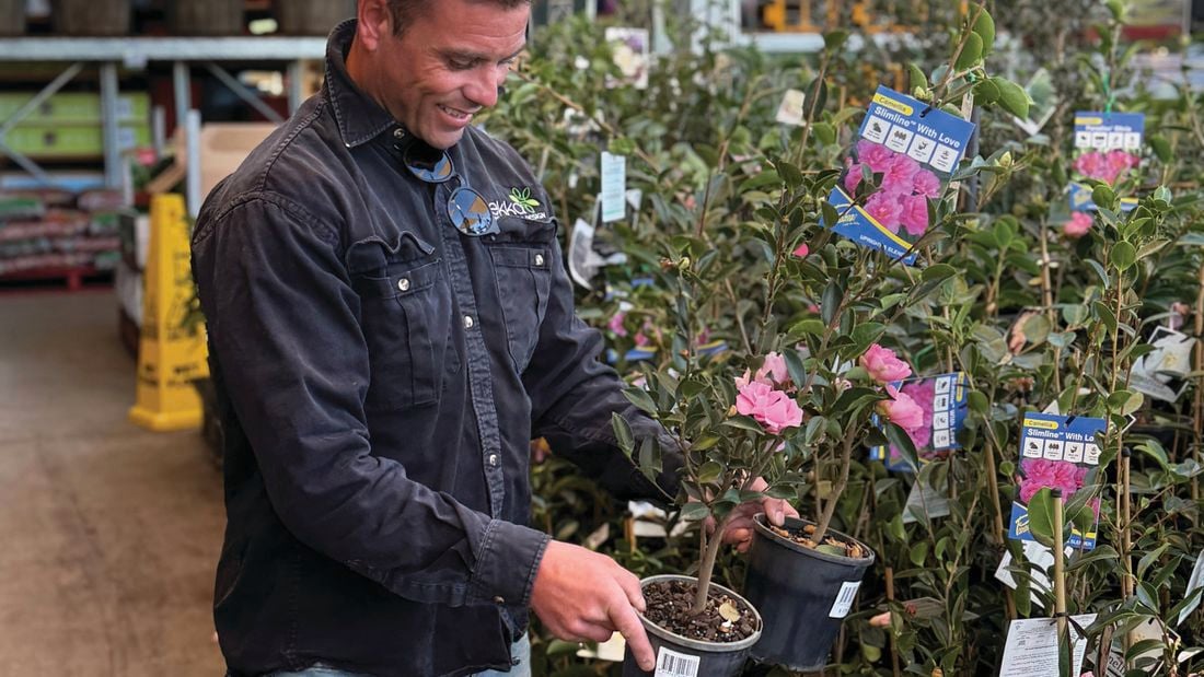 A tradesperson in a work shirt holds a plant with a pink flower in the garden section at Bunnings.]