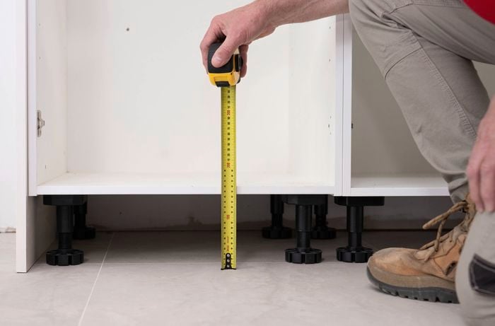 A person is measuring the height from the floor to the base of a kitchen cabinet using a tape measure to check whether the floor is even. 