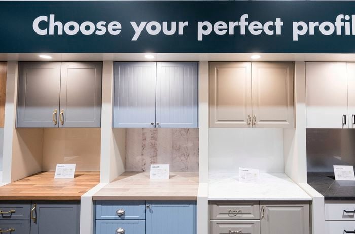 A green sign with wite writing on it - choose your perfect profile, above four kitchen displays with bench tops and cupboards in them.