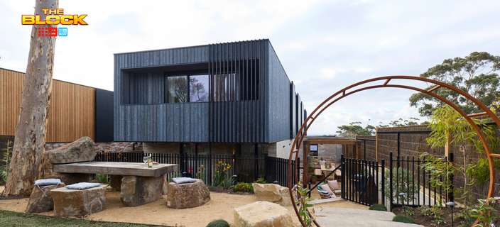 Modern two-storey home with dark cladding, landscaped garden, natural stone seating and a large circular rust-coloured metal sculpture.