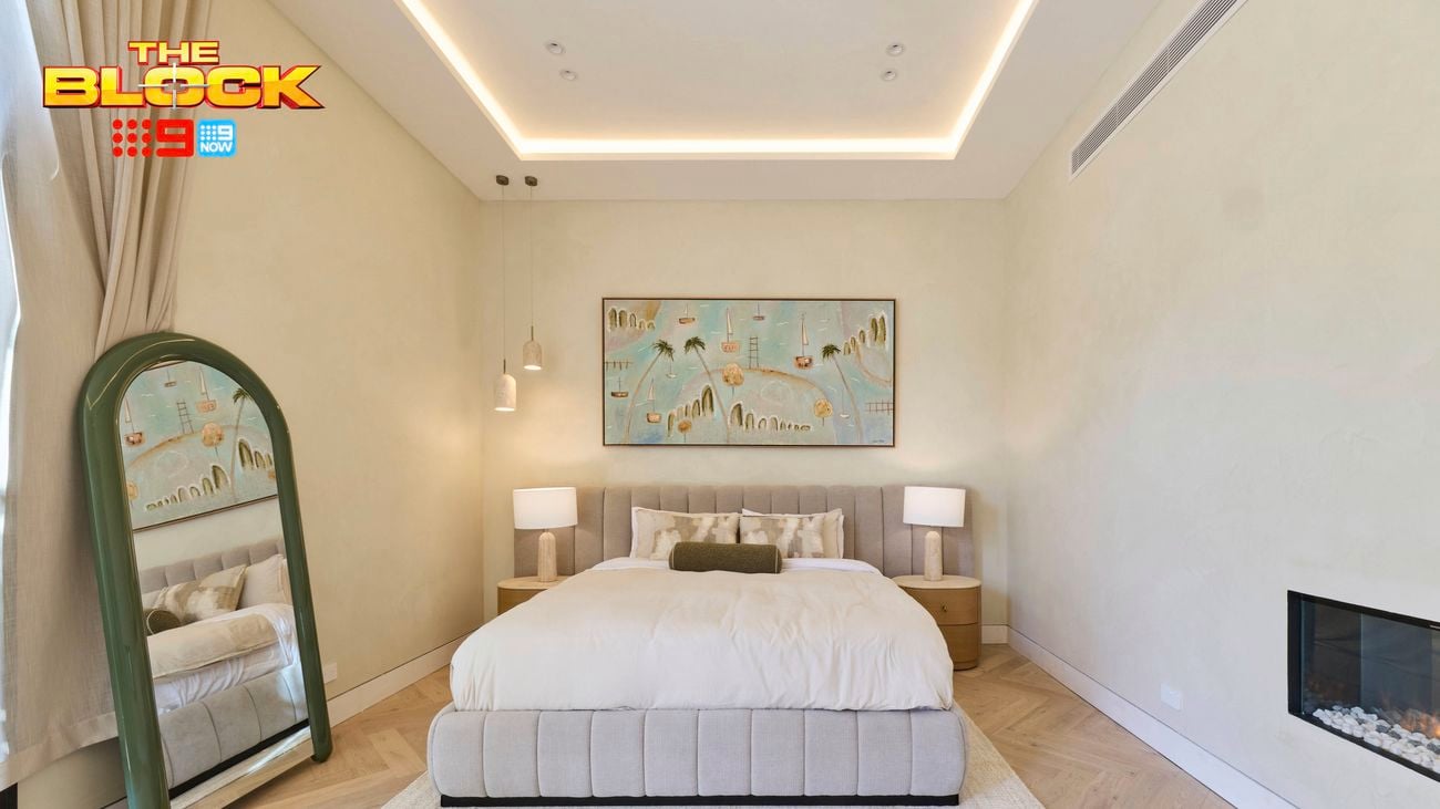Neutral bedroom with an upholstered bed, green arched floor mirror, bedside lamps, framed artwork, herringbone timber flooring and a built-in fireplace.
