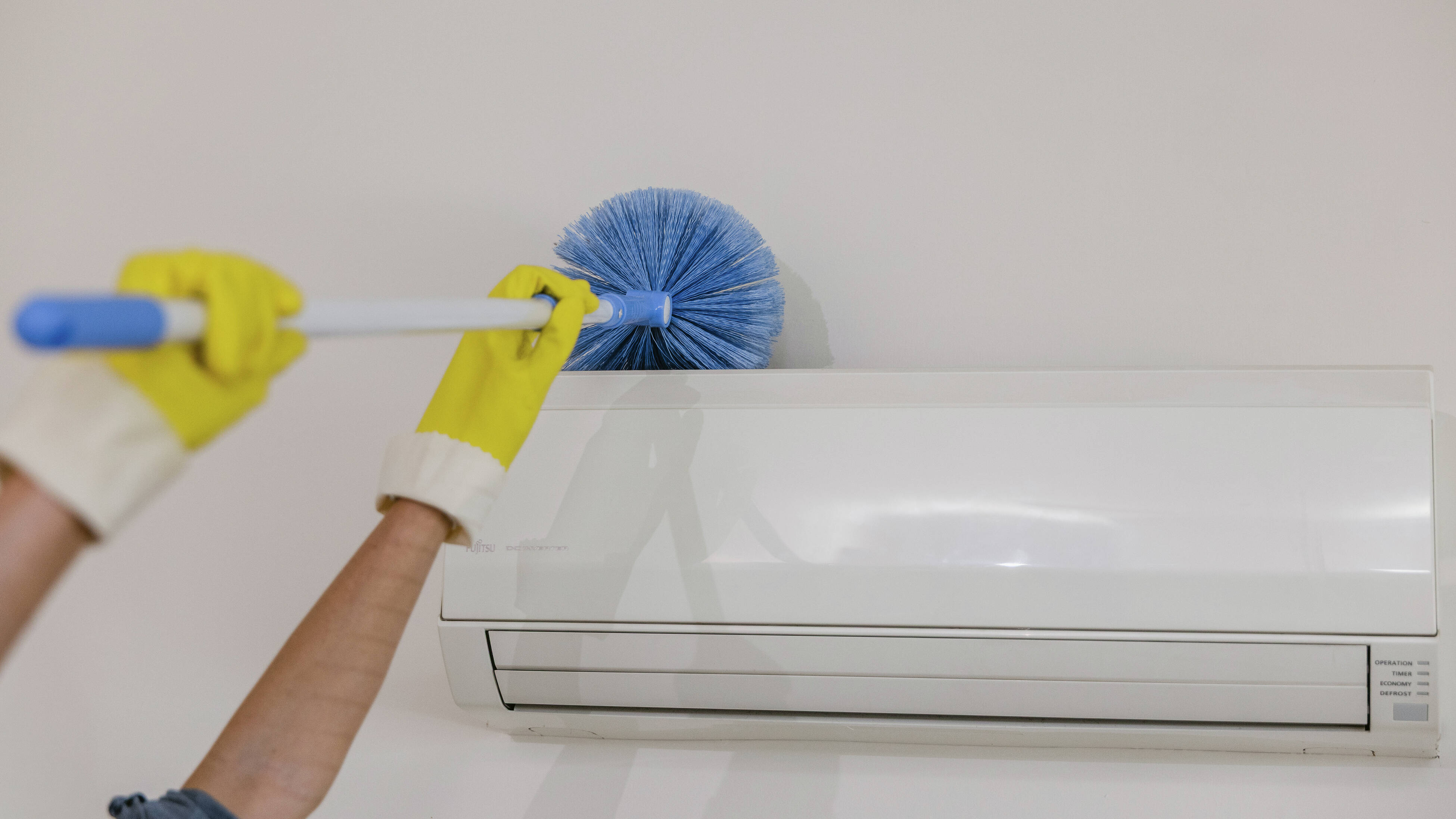 Gloved hands dusting a wall-mounted air conditioning unit with a blue microfiber duster.