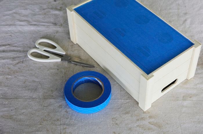 A wooden crate with painters tape covering the bottom of it. A pair of scissors and a roll of tape sit next to it