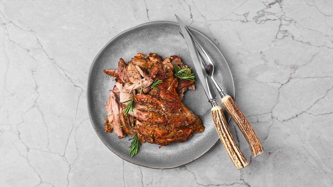 Carved lamb spit roast garnished with rosemary on a grey platter with a serving fork and knife.