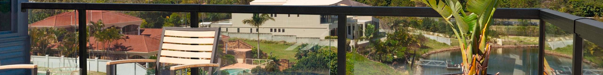 Balcony with outdoor furniture setting with a view of a modern house overlooking greenery and houses. Encased by a black aluminium balustrade with glass panels.