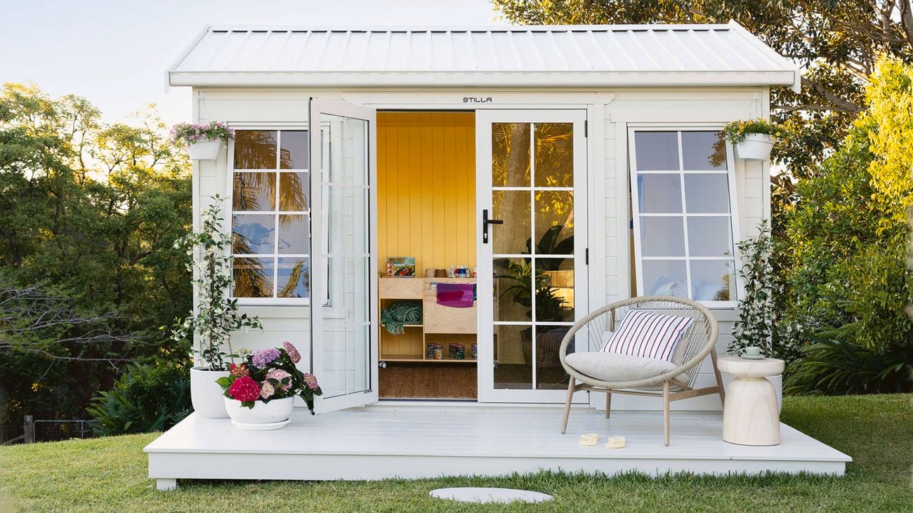 A front view of a large timber shed painted white with a front deck featuring hanging baskets, pot plants and a chair sits in a garden setting. One open French door shows the yellow interior and a timber cabinet on castors.