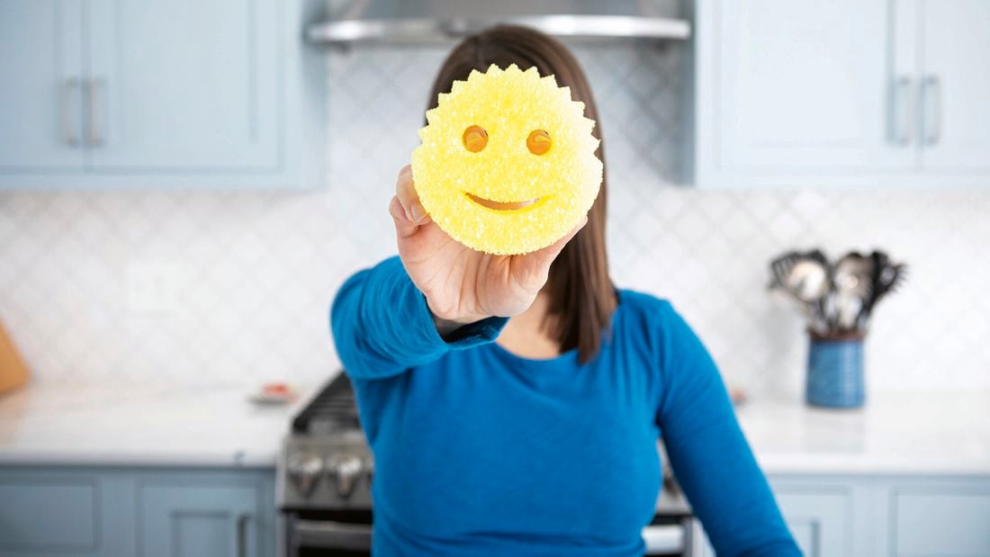 A woman holding a yellow Scrub Daddy sponge in front of her face, with a blue kitchen in the background.