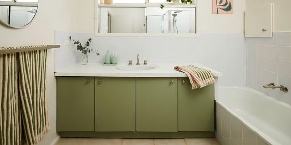 Bright bathroom with a green vanity cabinet and white benchtop, featuring a central sink beneath a wide mirror, a bathtub to the side, white tiled walls, striped towel on a rack, and potted plants along the windowsill for natural light and greenery.