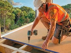 A construction worker in a hard hat and high-visibility vest kneels on timber sub flooring, using a power drill to install a floor panel inside a house overlooking the coast.