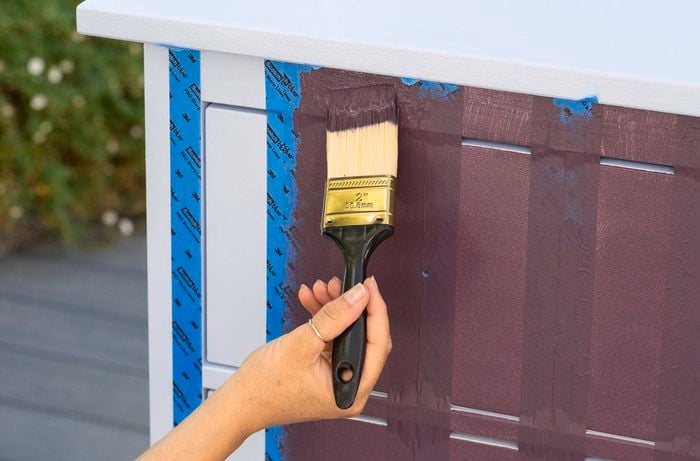 Paintbrush applying burgundy paint between vertical strips of painter’s tape on a cabinet drawer.