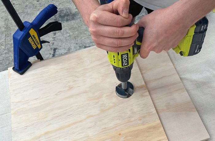 Hands using a cordless drill with a hole‑saw bit to cut a hole in a sheet of plywood clamped to a workbench.
