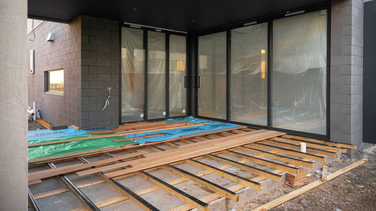 Partially completed outdoor deck area of a modern building under construction, featuring black-framed sliding glass doors, dark brick walls, timber decking boards being installed, and construction materials laid across the deck frame.