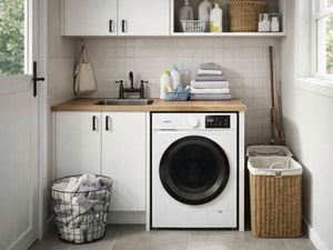 Laundry room with front‑loading washing machine under a wooden benchtop, white cabinetry, black tap and sink, folded towels, cleaning products, wire basket of laundry, and wicker hamper beside a window.