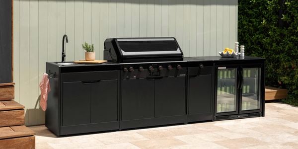 A black outdoor kitchen with a barbecue, sink and bar fridge in it, sitting on paving outside.