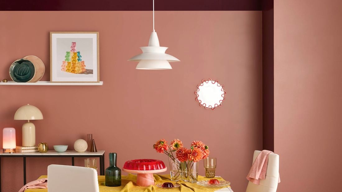 Modern dining room with two‑tone pink and plum walls and a white pendant light. A wall shelf holds framed art and ceramics, a dining table and chairs with a mustard tablecloth, glass vases and a red cake stand.