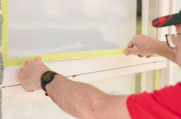A man applies yellow masking tape to the window exterior to prevent paint bleeding.